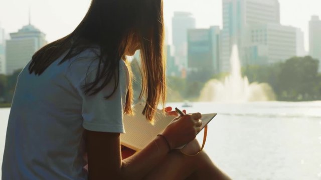 Young woman is making notes in her diary sitting in city park on the pond bank with fountain at sunset, back view. Woman is writing morning pages, 