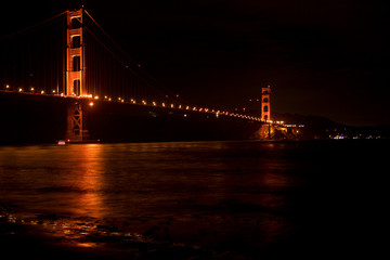 Obraz premium SAN FRANCISCO, CALIFORNIA, UNITED STATES - NOV 25th, 2018: Golden Gate Bridge as seen from Fort Point overlook is glowing in the night with star trails in the sky behind it