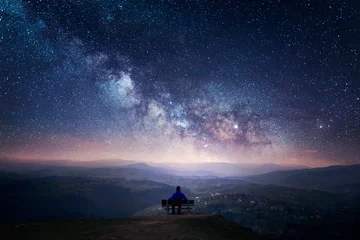  A man sitting on a bench staring at a starry sky with a Milky Way and a mountain landscape © PiotrKaluza