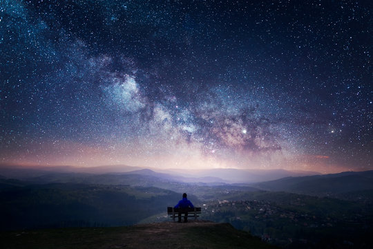 A man sitting on a bench staring at a starry sky with a Milky Way and a mountain landscape