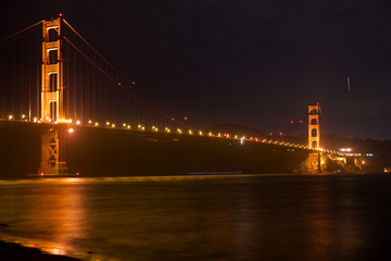 Fototapeta premium SAN FRANCISCO, CALIFORNIA, UNITED STATES - NOV 25th, 2018: Golden Gate Bridge as seen from Fort Point overlook is glowing in the night with star trails in the sky behind it