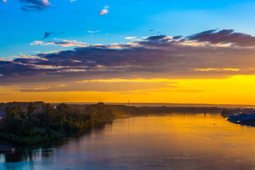 Bright colorful orange sunset over the river, dense dark forest on the shore, railway bridge across the river, reflection of the yellow blue sky in the water. Belaya river, Ufa, Bashkortostan, Russia.