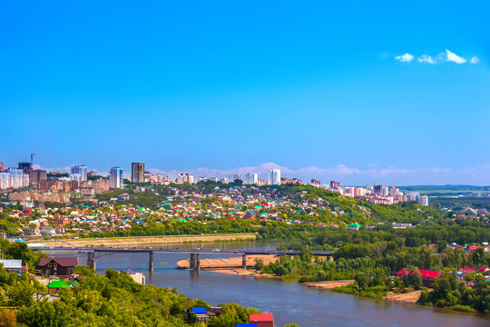 White River in the city center on the background of private houses with colorful roofs on a slope with green trees and modern high-rise buildings. Belsky Bridge, Ufa, Bashkortostan, Russia.