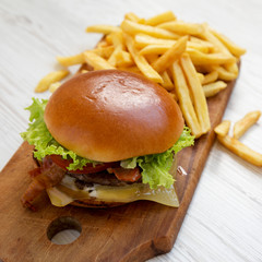 Hamburger with lettuce, cheese and bacon on a rustic wooden board on a white wooden table, side view. Close-up.
