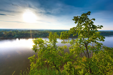 A large cliff overgrown with green grass and trees dominate over the White River in the evening at dusk during sunset under a clear blue sky. Rock Hanging Stone, Ufa, Bashkortostan, Russia.