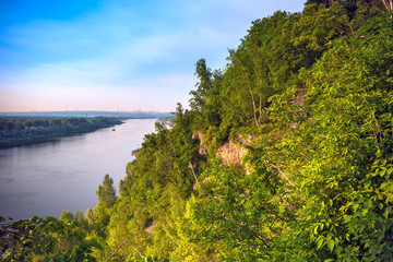 A large cliff overgrown with green grass and trees dominate over the White River in the evening at dusk during sunset under a clear blue sky. Rock Hanging Stone, Ufa, Bashkortostan, Russia.