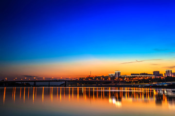 Belsky bridge over the river after sunset at night in the bright orange lights of the city with reflection in the water and a clean colorful blue sky at dusk. White river, Ufa, Bashkortostan, Russia.