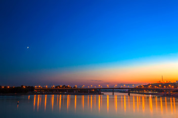 Obraz premium Belsky bridge over the river after sunset at night in the bright orange lights of the city with reflection in the water and a clean colorful blue sky at dusk. White river, Ufa, Bashkortostan, Russia.