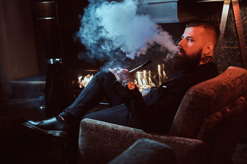 Bearded relaxed man is sitting on the armchair and smoking hookah near fireplace.