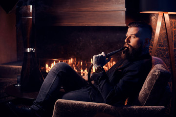 Groomed tattooed man is relaxing near fireplace in dark room and smoking hookah.