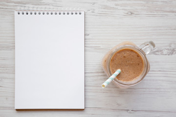 Banana apple smoothie in a glass jar, blank notepad over white wooden background, top view. Flat lay, from above, overhead.