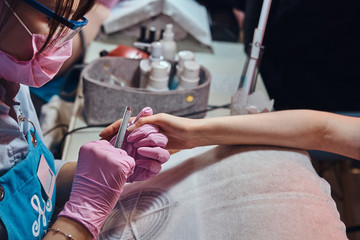 Talented dilligent manicurist in protective mask is working on customer nails with nail file.