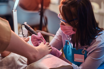 Young cheerful nail master is working on customer's toe nails at her own manicure studio.