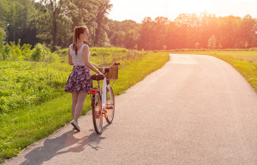 Girl with bike at the summer sunset on the road in the city park.Cycling down the street to work at summer sunset. Bicycle and ecology lifestyle concept.