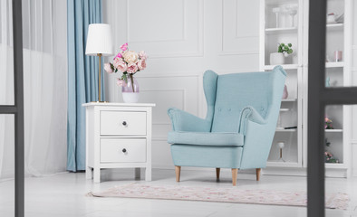 Light blue armchair and drawers cabinet with table lamp and flowers vase on it