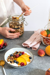 Female hands serving granola from jar, with greek yogurt fresh peach, blueberry, raspberry on the table - healthy breakfast