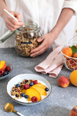 Female hands serving granola from jar, with greek yogurt fresh peach, blueberry, raspberry on the table - healthy breakfast