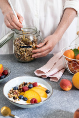 Female hands serving granola from jar, with greek yogurt fresh peach, blueberry, raspberry on the table - healthy breakfast