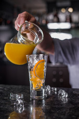 Pouring orange juice from glass carafe, on wooden table and bright background.