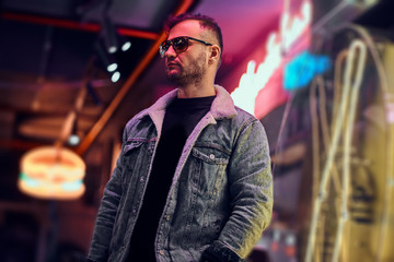 A bearded stylish male wearing a denim jacket and sunglasses looking away while standing in the night on the street. Illuminated signboards, neon, lights.