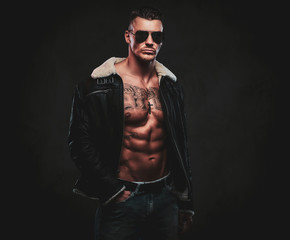 A macho stylish man in jeans jacket with dark glasses and stylish hair on a dark background.