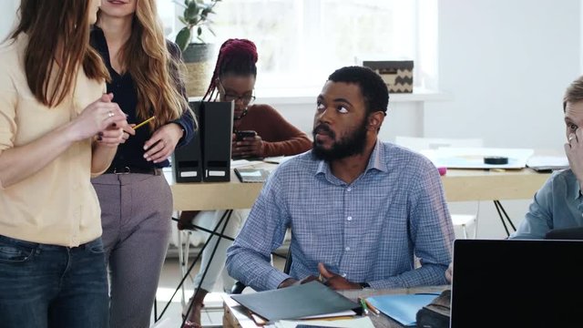 Young intelligent Caucasian business woman giving paperwork documents to upset African male employee at modern office.