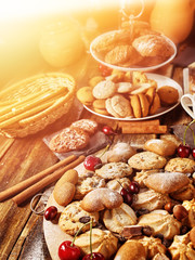 Oatmeal cookies and sand chocolate cake with cherry on cutting board on wooden table in rustic style served with cups and dessert spoons. Sun flare morning sunrays from window. Sunlight in kitchen.