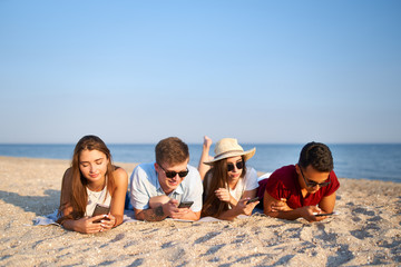 Group of millenials using smartphones laying together on beach towel near sea on summer sunset....