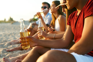 Group of friends having fun enjoying refreshing beverage and relaxing on the beach at sunset. Young...