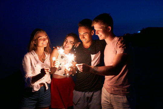 Friends walking, dancing and having fun during night party at the seaside with bengal sparkler lights in their hands. Young teenagers partying on the beach with fireworks. Slow motion steadycam shot.