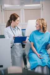 Obraz premium cheerful doctor holding clipboard while standing near patient sitting in armchair