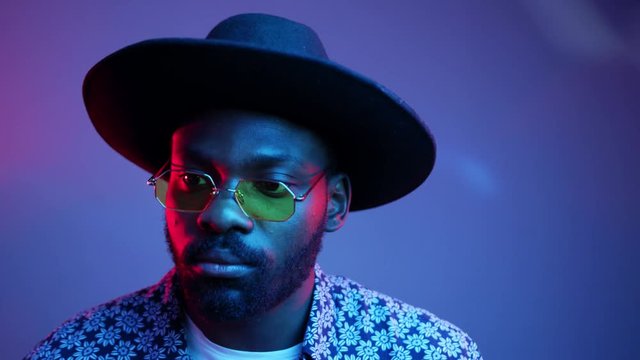 African American stylish man with hat in glasses smiling look at camera smile posing on the blue and violet neon light background attractive beard black blue boy bright cheerful slow motion