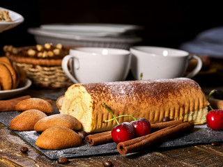 Oatmeal cookies and rolled cake on tier cake stand with cherry and nut on kitchen on wooden table in village style. Two cups of tea on wooden table. Delight pleasant taste. Limitations for diabetics.