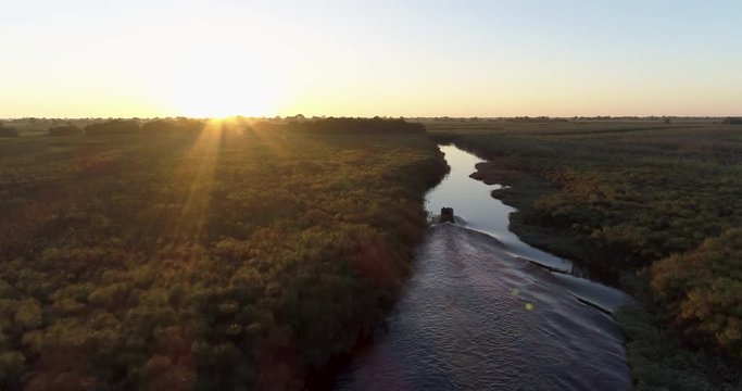 Aerial backlit view of a tourist boat making its way down one of the many curving waterways of the Okavango Delta at sunset