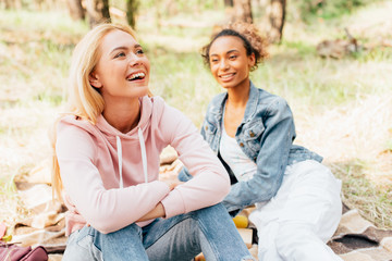 Fototapeta premium two laughing multiethnic friends sitting on plaid blanket in forest