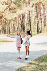 Fototapeta premium full length view of two smiling multicultural friends holding hands while skateboarding on penny boards on road