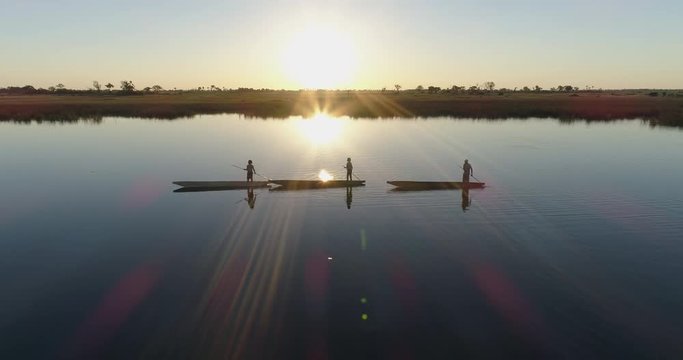 Aerial fly over view of three polers silhouetted against the setting sun rowing their Mokoros on the waterways of the Okavango Delta