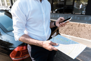 cropped view of businessman holding smartphone and map while standing near car