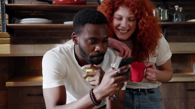 African American man eat apple with curly red woman use their smart phone couple happy smile talk at home at kitchen girlfriend internet browsing pay technology together date shopping slow motion