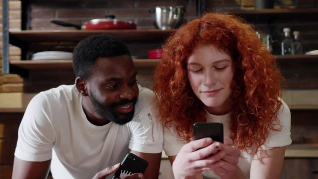 Close up african American man eat apple with curly red woman use their smart phone couple happy smile talk at home at kitchen girlfriend internet browsing technology together date shopping slow motion