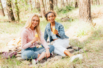 two multiethnic girls sitting on plaid blanket and holding bottles of orange juice