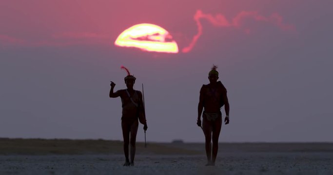 Two Bushman walking aross the Makgadikgadi Pans with the sun setting in the red sky behind them, Botswana