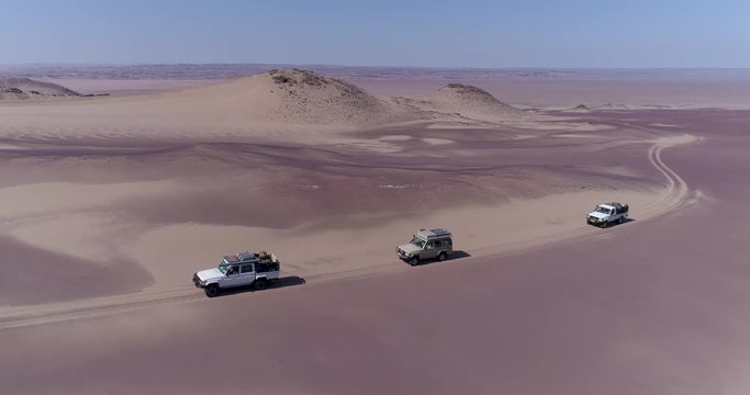 4K aerial zoom out view with beautiful scenery of a convoy of 4x4 vehicles driving through the sand dunes and mountains on the Skeleton Coast, Namib desert, Namibia