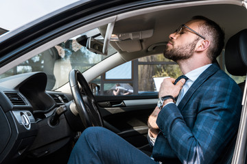 low angle view of bearded businessman in glasses touching tie while sitting in car