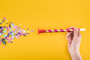 cropped view of woman holding party horn near confetti on yellow background