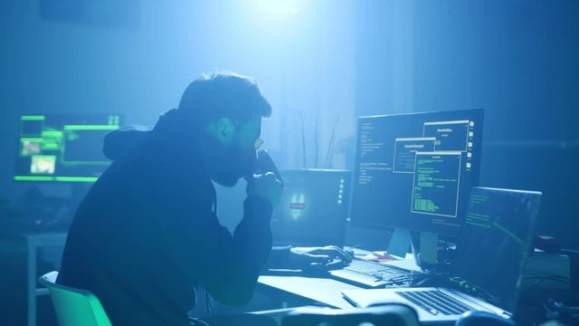 Group of internationally wanted hackers organising cyber malware attack on government at night. Back view of hooded hacker working on computer in dark hideout.