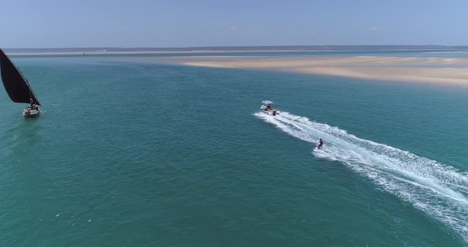 Aerial back view of a pretty young girl wakeboarding pulled by a speedboat on an beautiful scenic estuary with dhow sailing boats in the background