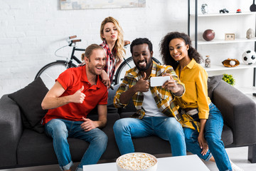 Excited happy multicultural men and women sitting on couch and taking selfie in living room