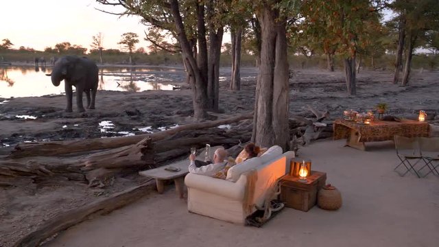 4K close-up panning view of tourists sitting at sundown with the evening fire lit, watching an elephant drinking at the waterhole in front of Khwai Private Reserve, Botswana