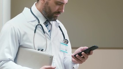 Up-section shot of male surgeon staying in hallway and reading research article on his mobile phone and going out